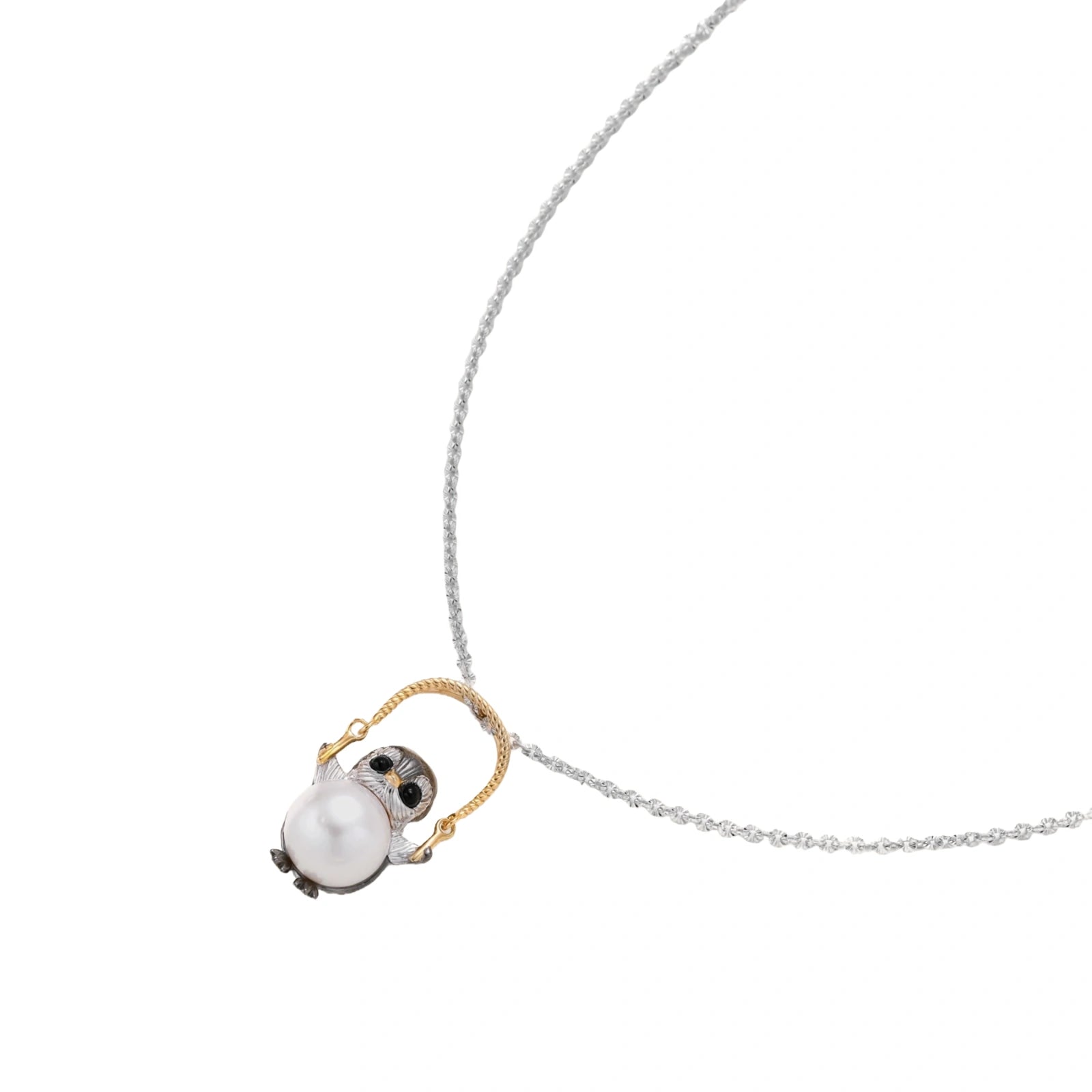 Whimsical owl pendant holding a white pearl on a gold swing, rhodium-plated silver chain on cream background.