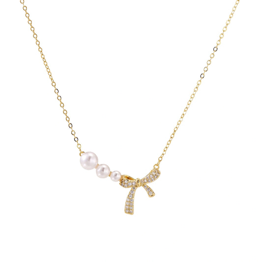 Delicate gold necklace with three white pearls and crystal-embellished bow pendant on cream background